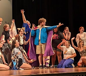 Take It From The Top: A group of performers energetically posing on stage, with one wearing a cape in the center.