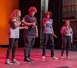 Take It From The Top: Four young people holding microphones on a lit stage.